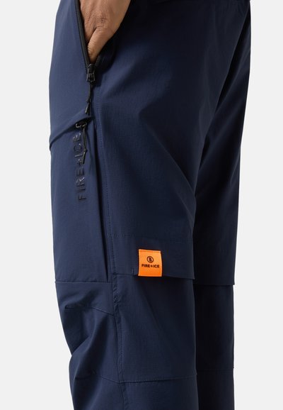 Zbliżenie na granatowe spodnie z zasuwaną kieszenią i pomarańczową metką z logo FIRE+ICE na boku, ręka częściowo włożona do kieszeni.
