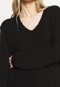 Schwarzer gestrickter Pullover mit V-Ausschnitt-Design, mit gerippter Textur und langen Ärmeln, der teilweise eine Hand in entspannter Position zeigt.