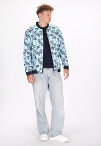 Giubbotto bomber azzurro cielo con stampa di palme, colletto e polsini a coste neri, indossato sopra una camicia blu navy, abbinato a jeans chiari e sneakers bianche.