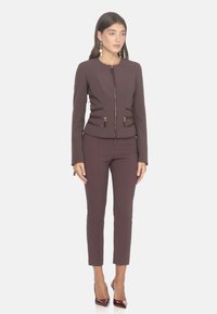 Giacca aderente color malva con chiusura a zip, due tasche anteriori e dettagli nelle cuciture; abbinata a pantaloni slim-fit coordinati e tacchi borgogna.