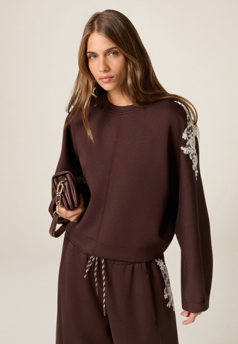Bruine sweatshirt met lange mouwen, voorzien van witte bloemenborduursels op de schouder. Gekozen met bijpassende joggingsbroek en een donkerbruin handtas.