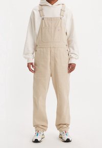 Peto de pana beige con piernas anchas, tirantes ajustables y bolsillo delantero con un pequeño parche de logo. Se lleva sobre una sudadera blanca con capucha y zapatillas.