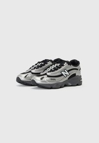 Silver athletic shoes with a textured upper, featuring black accents and laces, a curved design, and a chunky rubber sole.