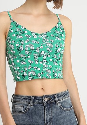 Kvinde iført en grøn blomsterprint crop top med tynde stropper og højtaljede blå jeans foran en ensfarvet baggrund.