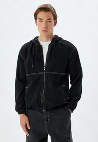 Zwarte hoodie met een ritssluiting aan de voorkant, elastische manchetten en een twee-kleurig ontwerp met witte stiksels en zijzakken. Gemaakt van denimstof.