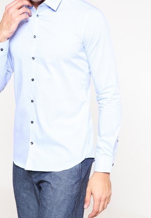 Mann trägt ein hellblaues Hemd mit langen Ärmeln und dunkelblaue Hose, posiert mit einer Hand in der Nähe seines Gesichts.