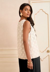 Mujer con cabello largo y oscuro lleva una camiseta blanca sin mangas con lunares en relieve, pantalones negros, pendientes geométricos y una pulsera de oro, de pie en el interior.