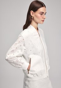 Giacca bomber bianca con motivi floreali ricamati, design ad occhiello, polsini e orlo a costine, e una zip frontale con tasca rotonda.