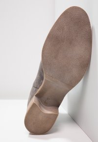 Botín de ante gris con suela texturizada, con un tacón ligeramente curvado. El interior muestra un forro liso beige y una marca mínima.