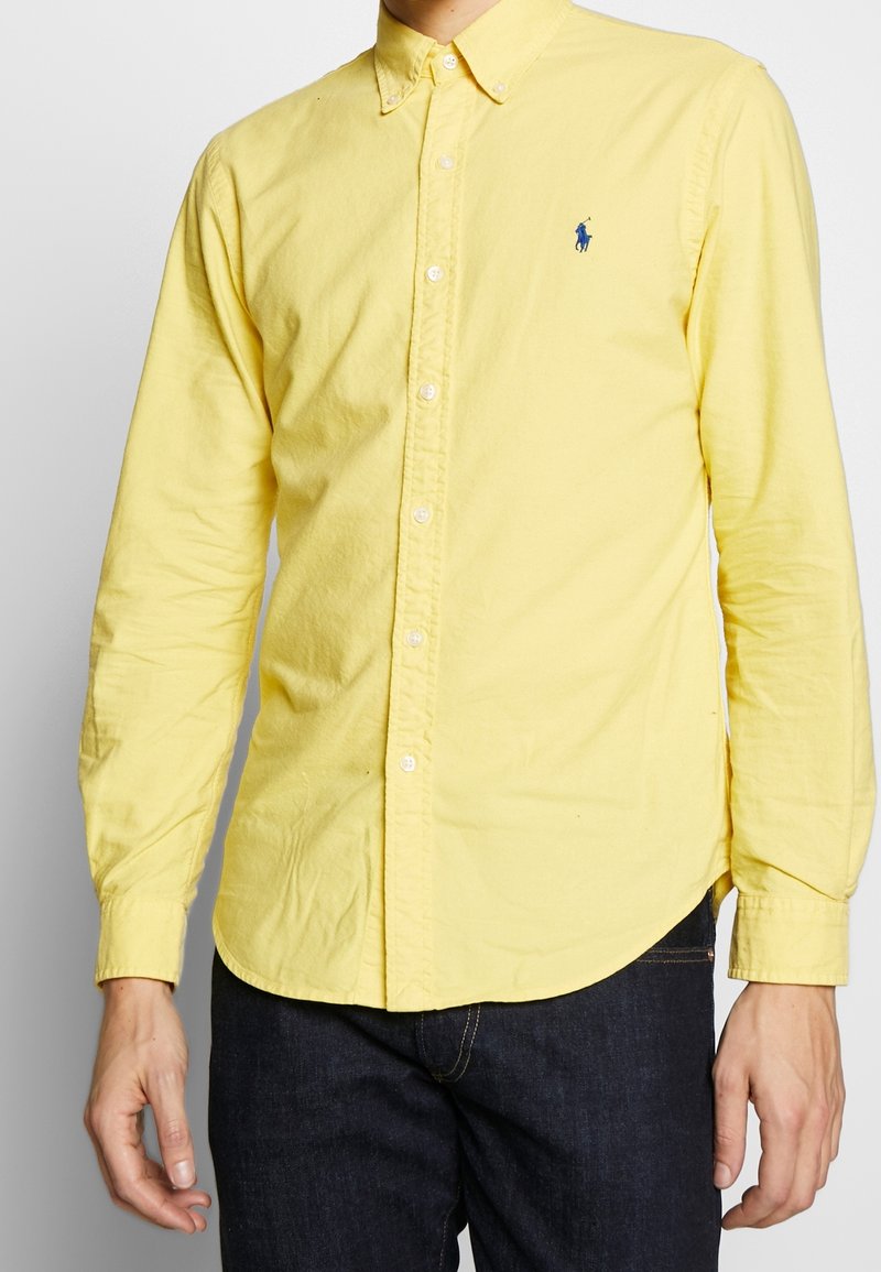 Żółta koszula na guziki wykonana z bawełny, z zaokrąglonym dołem, długimi rękawami i małym niebieskim logo na lewej piersi.