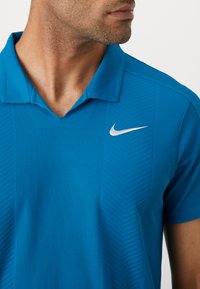 Kék polo póló textúrált anyagból, függőleges csíkokkal. A bal felső részen egy jellegzetes, ezüst Nike logó található.