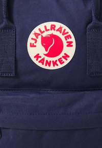 Mochila Kånken da Fjällräven em azul marinho, destacando um logotipo redondo com a silhueta de uma raposa vermelha e texto em rosa sobre um fundo creme.