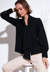 Black blouse with a bow tie at the neck, long sleeves, and puffed cuffs. Soft fabric with a smooth texture. Paired with light-colored pants.
