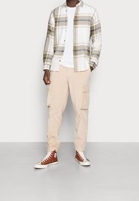 Man som bär beige kordjacka cargo-byxor, bruna högskor, vit t-shirt och en skjorta med knappar i beige, vitt och grått rutmönster.