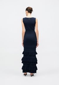 Γυναίκα με μαλλιά σε κότσο, που φοράει αμάνικο μακρύ σκούρο μπλε φόρεμα με στρώσεις βολάν στο πίσω μέρος και καφέ σανδάλια με τακούνι, στέκεται σε λευκό φόντο.