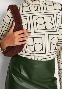 Maglia a maniche lunghe di colore crema con motivi geometrici neri; abbinata a una gonna in ecopelle verde scuro; tiene in mano una borsa a spalla marrone pelosa.