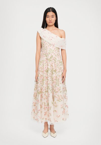 Kvinde iført en hvid, one-shoulder, blomstrede kjole med pink og grøn broderi, parret med creme farvede spidse sko, stående foran en hvid baggrund.