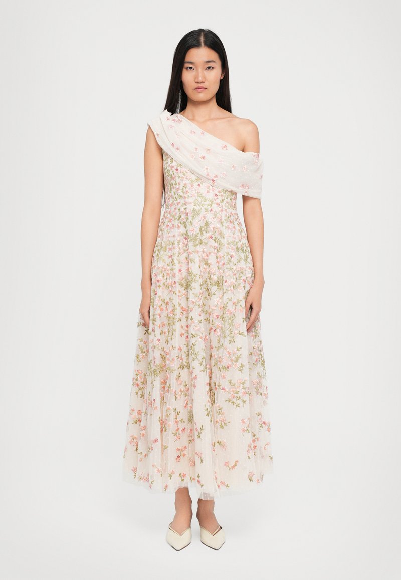 Vrouw in een witte, een-schouder, bloemenjurk met roze en groene borduursels, gecombineerd met crèmekleurige, spitse schoenen, staande tegen een witte achtergrond.