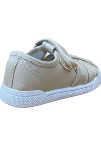 Zapatilla de lona en beige con banda elástica, suela de goma blanca, textura antideslizante y detalles de costura en la parte superior. Logo de la marca en el talón.