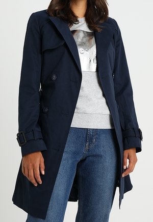 Frau trägt einen marineblauen, zweireihigen Trenchcoat über einem hellgrauen Sweatshirt und blauen Jeans vor weißem Hintergrund.