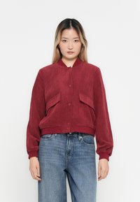 Woman wearing a ribbed dark red bomber jacket with front pockets and blue high-waisted jeans, standing against a plain white background.
