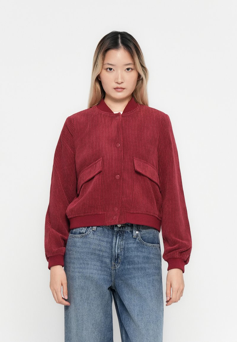 Woman wearing a ribbed dark red bomber jacket with front pockets and blue high-waisted jeans, standing against a plain white background.