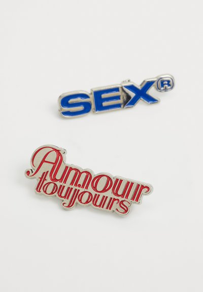 Metalli enamel pinni: "SEX" sinisessä hopeareunalla ja "Amour toujours" punaisena pyörteillä, molemmat kiiltävällä pinnalla.