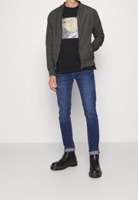 Olivgrön bomberjacka över en svart grafisk sweatshirt, kombinerad med blå jeans. Modellen har på sig svarta Chelsea-stövlar med gul söm.