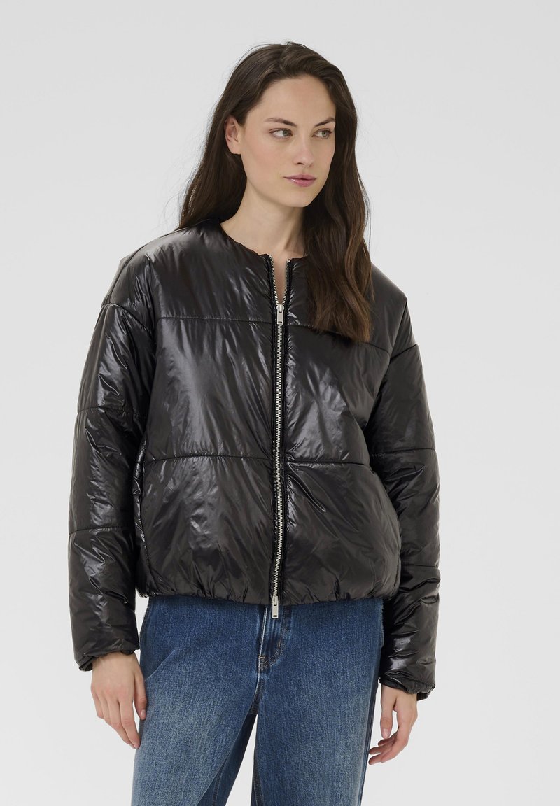 Chaqueta acolchada negra con acabado brillante, cuello redondo, cierre de cremallera y diseño de paneles cosidos, usada sobre vaqueros de mezclilla.