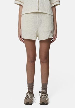 Personne portant un short en maille texturée crème, un haut assorti à manches courtes, des chaussettes brunes et des baskets beiges, debout sur un fond blanc.