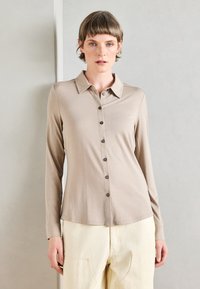 Bež sako sa dugim rukavima od mekog materijala, s klasičnom kragnom i tamnim dugmadi, nosi se uz kreme boje hlače.