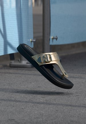 Μαύρο σανδάλι με χρυσό λουράκι και αγκράφα που αιωρείται πάνω από την άσφαλτο κοντά σε μπλε υφασμάτινα πάνελ.