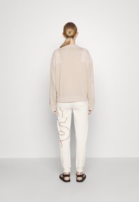 Beige Sweatshirt mit lockerem Schnitt und beigefarbenen Nähten, kombiniert mit cremefarbenen Hosen mit braunem Logodesign. Getragen mit schwarzen Sandalen.