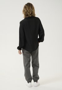 Camisa negra de manga larga con tejido texturizado y cuello con volantes, combinada con pantalones grises ajustados y zapatillas blancas. Vista trasera visible.