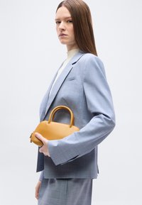 Γυναίκα με μακριά καστανά μαλλιά που φοράει ανοιχτό μπλε κοστούμι κρατά μικρή ταμπά δερμάτινη τσάντα σε απλό λευκό φόντο.