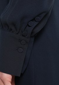 Námořnicky modrá blůza s měkkou tkaninou, nafouklými rukávy a dekorativními knoflíkovými detaily na manžetách pro přidanou strukturu.