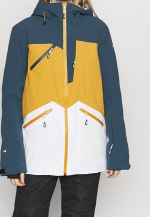 Personne portant une veste d'extérieur tricolore avec des parties bleu sarcelle, jaune moutarde et blanc, et plusieurs poches zippées.