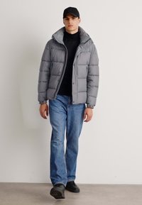 Gray puffer jacket with a hood, featuring a quilted design and front buttons, paired with dark jeans and black shoes. Simple cap completes the look.