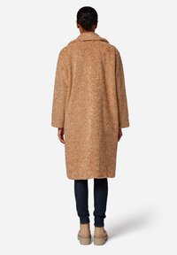 Oversized jas in cognackleur gemaakt van gestructureerde stof, met een brede kraag en mouwen over de volle lengte, gedragen over donkere spijkerbroeken en beige laarzen.