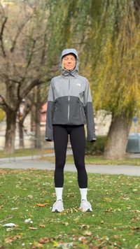 Jachetă gri și neagră impermeabilă cu fermoar și glugă, asortată cu colanți negri și șosete și pantofi sport albi, stând pe iarbă.