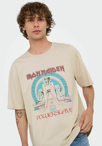 Camiseta de algodón beige con un estampado gráfico de estatuas inspiradas en Egipto y el texto "Iron Maiden Powerslave" en letras rojas en negrita.
