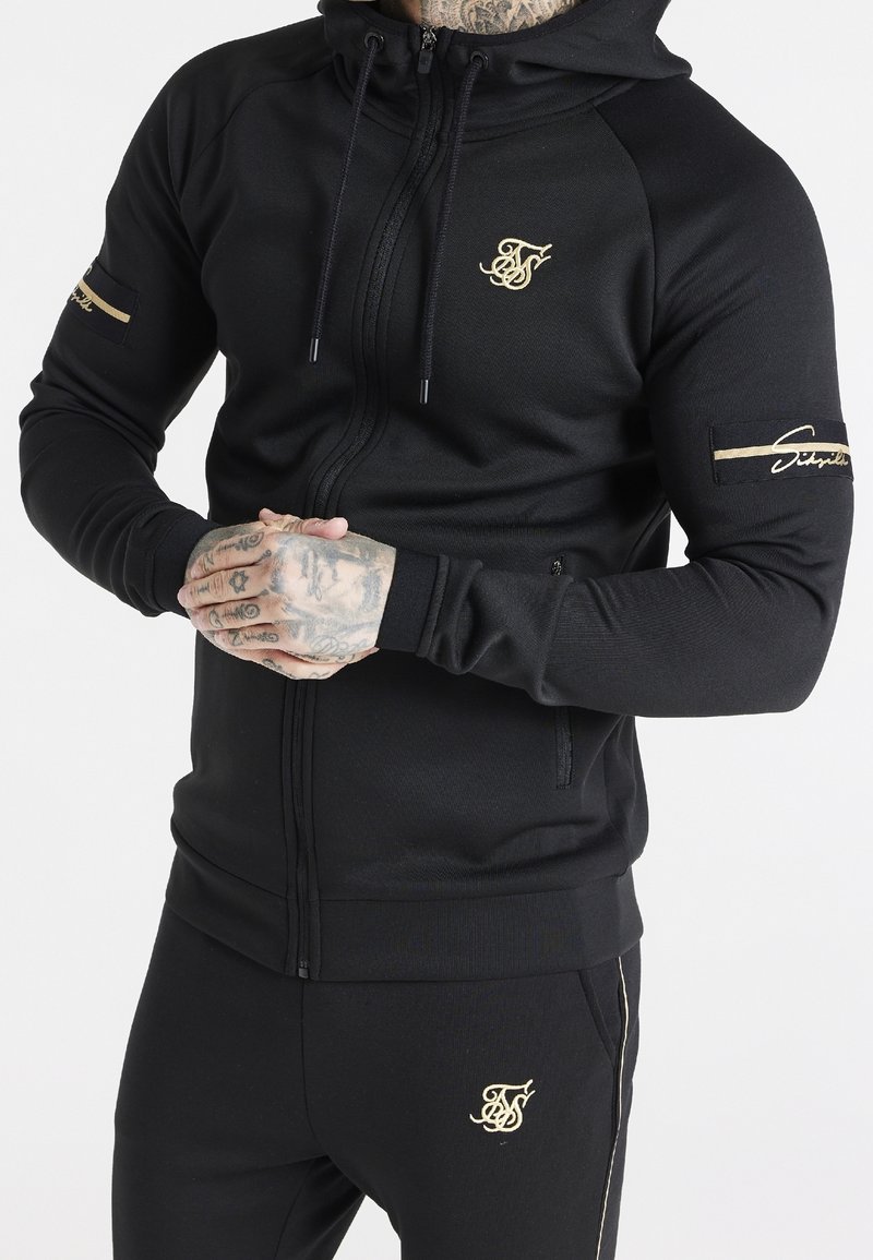 Homme portant un sweat à capuche noir zippé et un pantalon assorti avec des logos brodés en or, mains jointes, montrant des doigts et poignets tatoués.