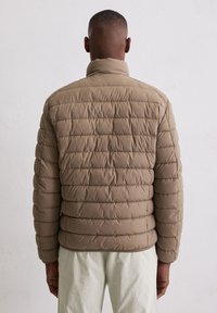 Beige donsjack met doorgestikt ontwerp, hoge kraag en elastische manchetten, gedragen over lichtgekleurde broek. Gladde textuur, minimale details.