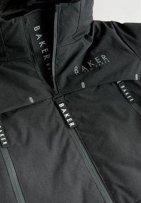Schwarze Zip-Jacke aus glattem, mattem Stoff mit hohem Kragen und Logo-Akzenten. Zwei Reißverschlüsse mit gebrandeten Zippern und sichtbaren Fake-Taschen.