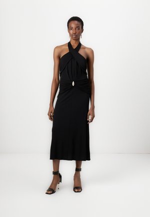 Black halter dress with gathered waist detail and a decorative gold accent. Smooth fabric drapes to knee length. Black high-heel sandals.
