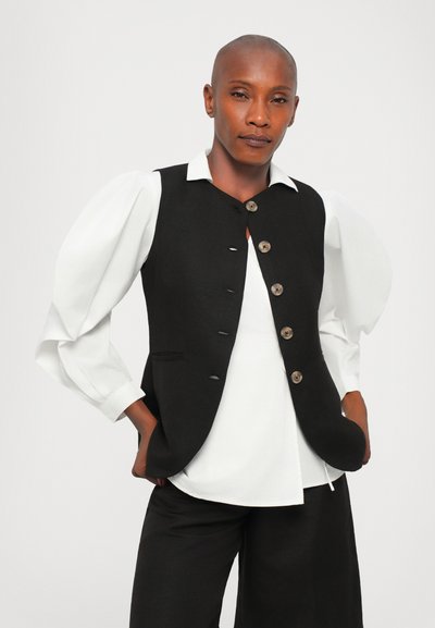 Black sleeveless vest with five buttons, worn over a white shirt with puffed sleeves, paired with black pants, on a neutral background.