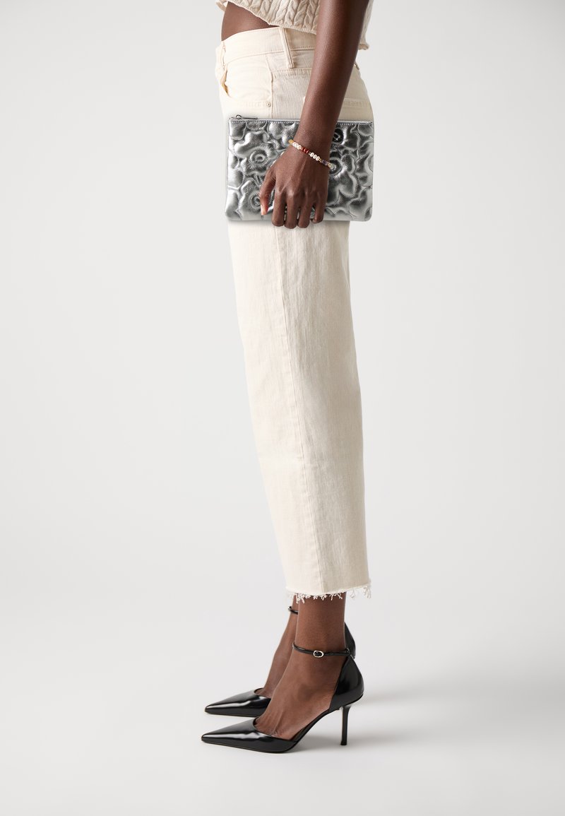 Clutch plateado texturizado sostenido con una mano, pantalones de longitud ankle de color claro, ligeramente desgastados, y tacones de charol negros de punta afilada.