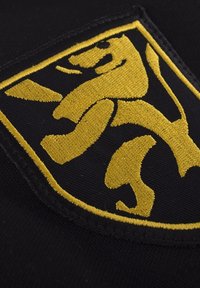 Zwarte stoffen patch met een geborduurd geel leeuwsymbool. Het ontwerp bevat gebogen elementen en een rand rondom de patch.