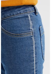 Blå denimjeans med en närbild av bakfickan och sid sömmen, som har gul söm och ett kontrasterande vitt sömdetalj.