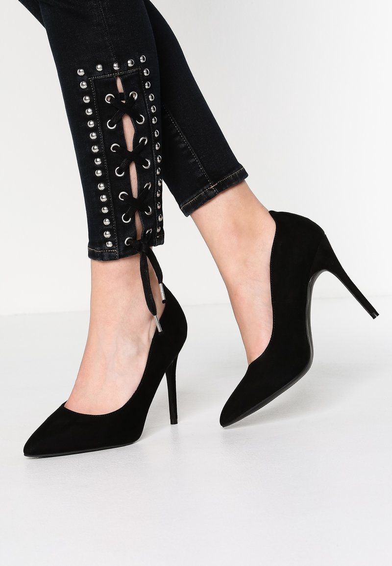 Escarpins noirs en cuir suédé à bouts pointus, portés avec un jean skinny noir présentant des détails lacés et cloutés aux chevilles.